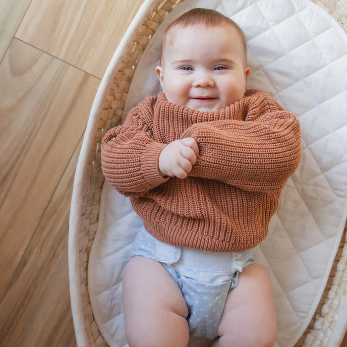 Baby wearing a brown knitted sweater in a crib with wooden floor background