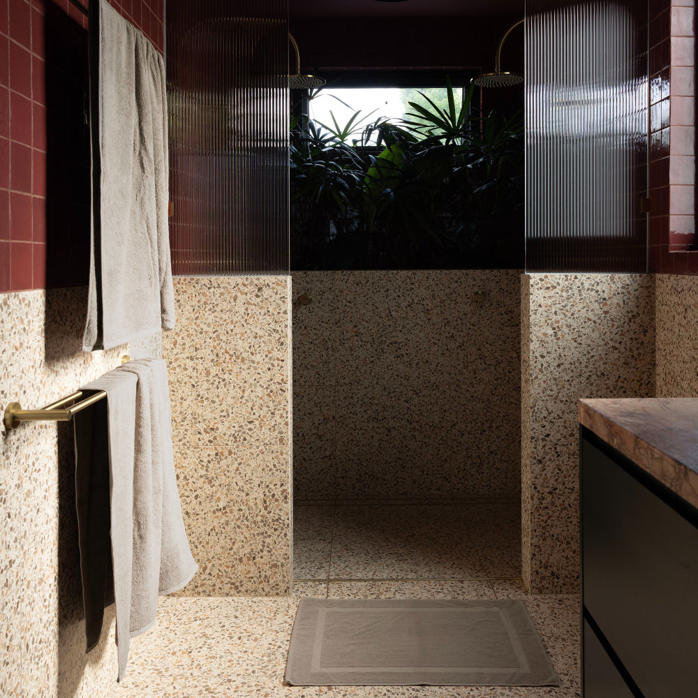The Bambury bath mat in latte is pictured on the floor in front of the shower with the bath towel and bath sheet in latte also hanging on the towel rail.