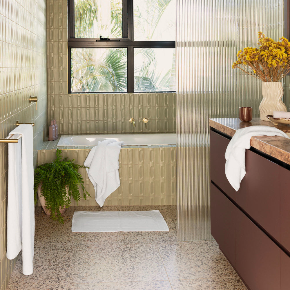 The Bambury Chateau bath mat white is placed on the bathroom floor in front of the bathtub with the rest of the white Chateau range hanging and placed around the rest of the bathroom.