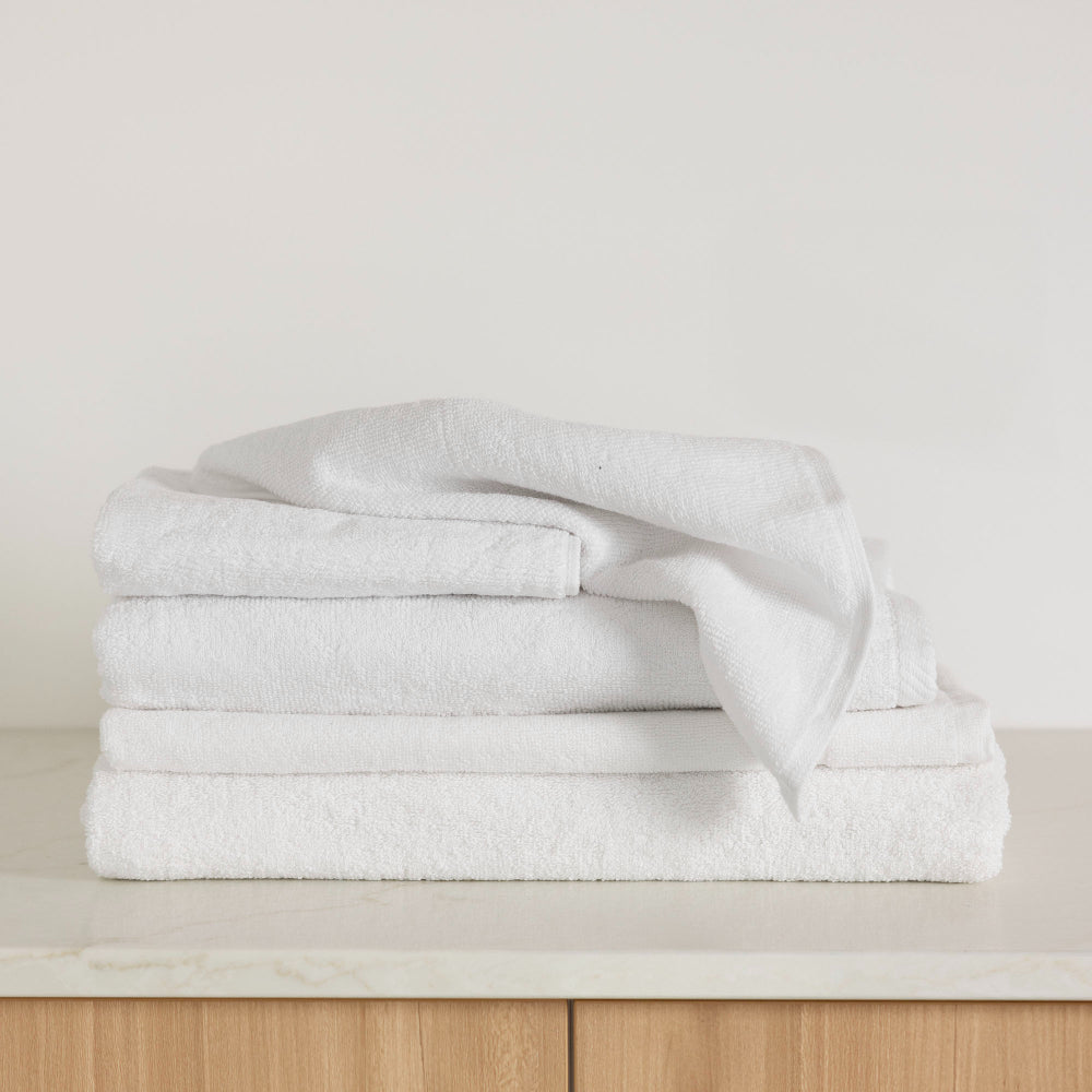 The Bambury Chateau bath mat, hand towel, face washer, bath towel and bath sheet in white is folded together on a bathroom vanity clean and ready to use.