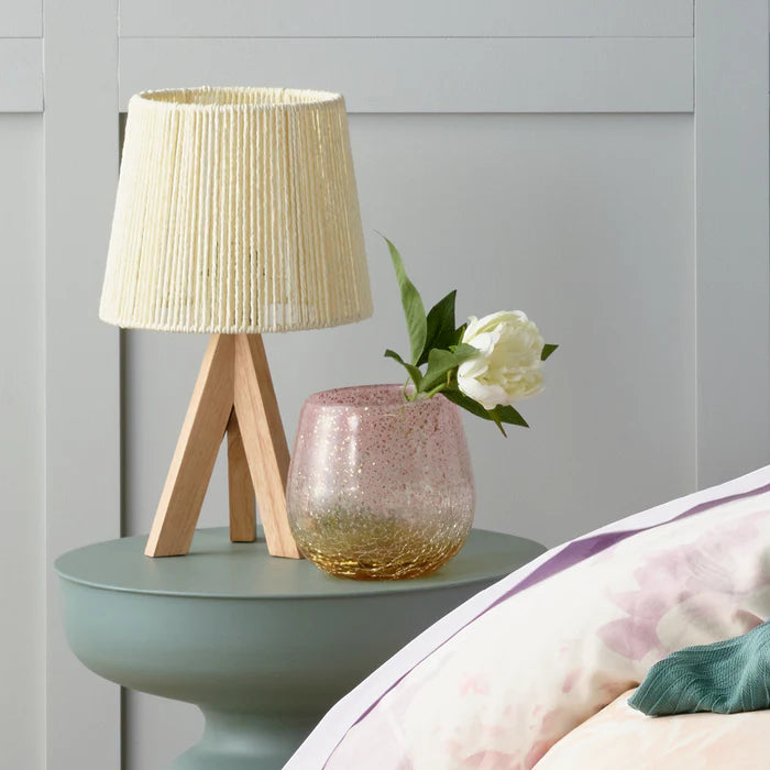 Small lamp with a textured shade on a bedside table next to a vase with a white flower.