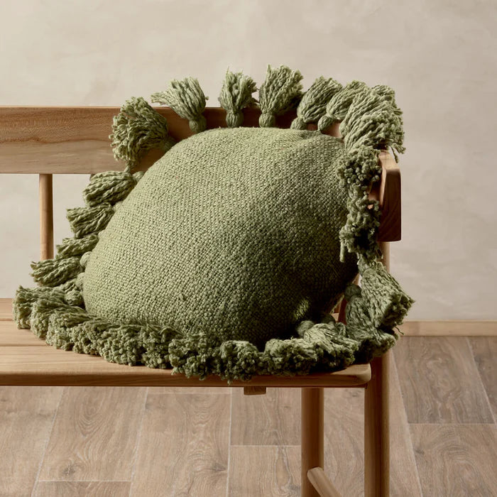 Green knitted cushion with tassels on a wooden chair against a neutral background