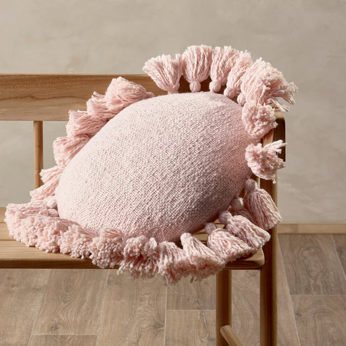 Pink cushion with tassels on a wooden chair against a neutral wall.