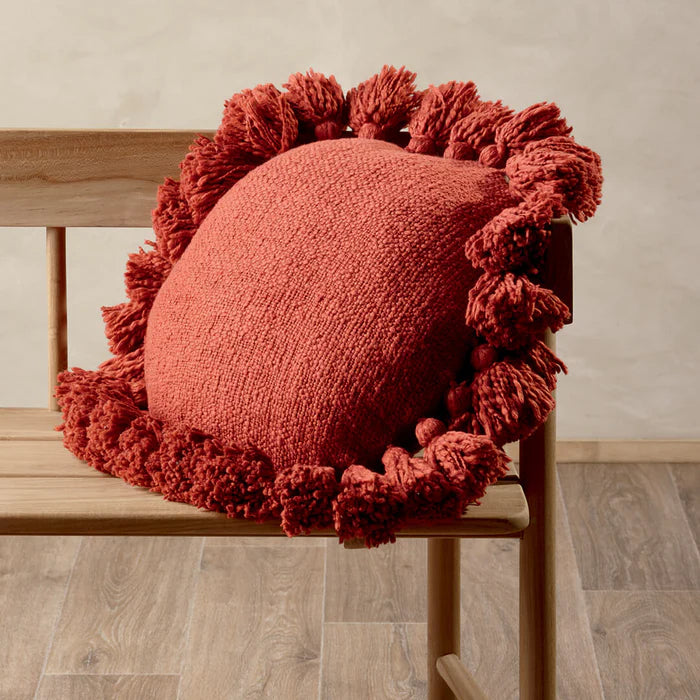 Red textured cushion with pom-pom trim on a wooden chair against a neutral wall.