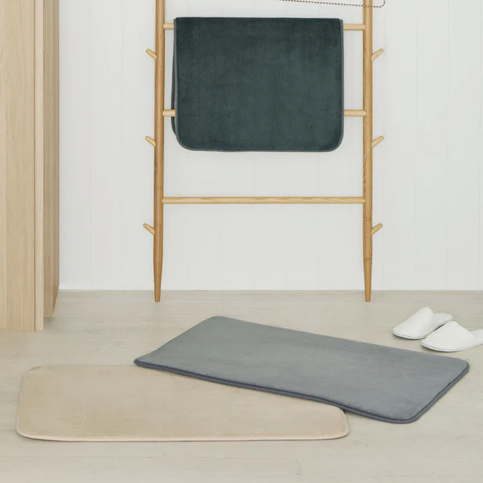 Two bath mats, one gray and one beige, on a wooden floor with a towel rack in the background.