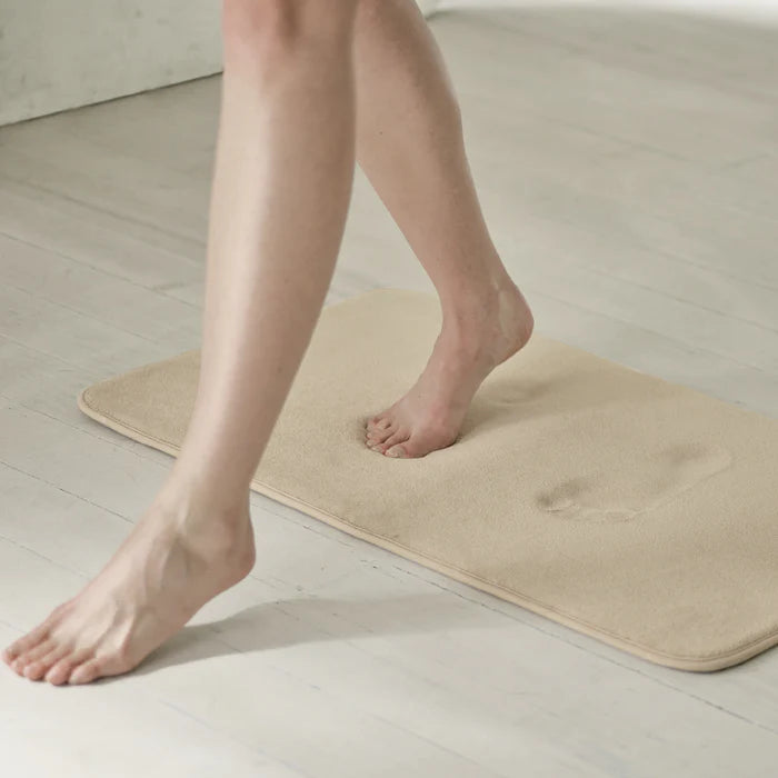 Person stepping onto a beige bath mat on a light wooden floor.