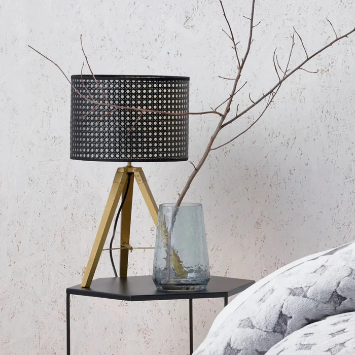 Table lamp with black woven shade and gold base on a small table against a textured wall.