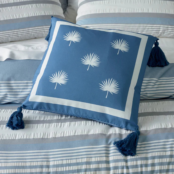 Blue pillow with white palm leaf patterns and tassels on a striped bed.