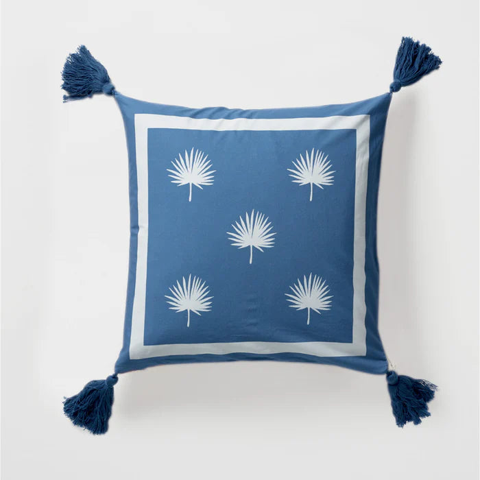 Blue pillow with white palm leaf patterns and tassels on a light gray background
