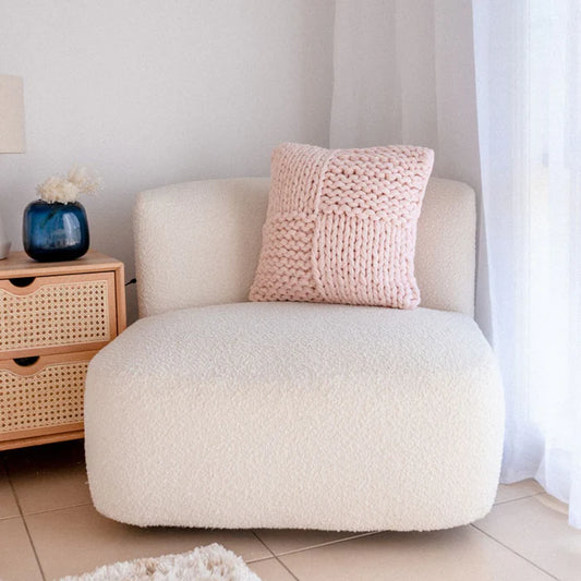White armchair with a pink knitted pillow in a room with a wooden side table and white curtains.