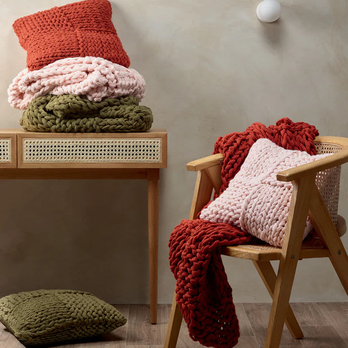 Knitted blankets and pillows in various colors on a wooden chair and table against a neutral wall.