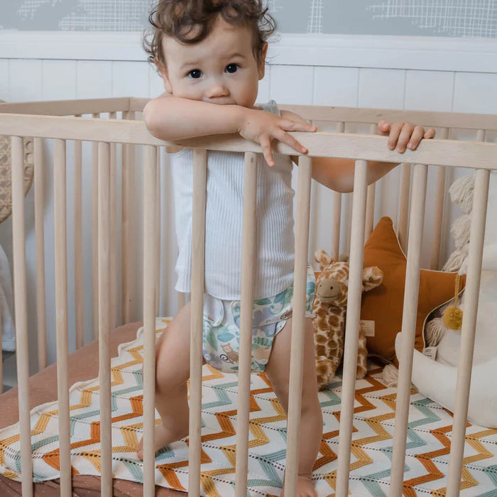 Child standing in a crib with a colorful blanket and stuffed giraffe toy.