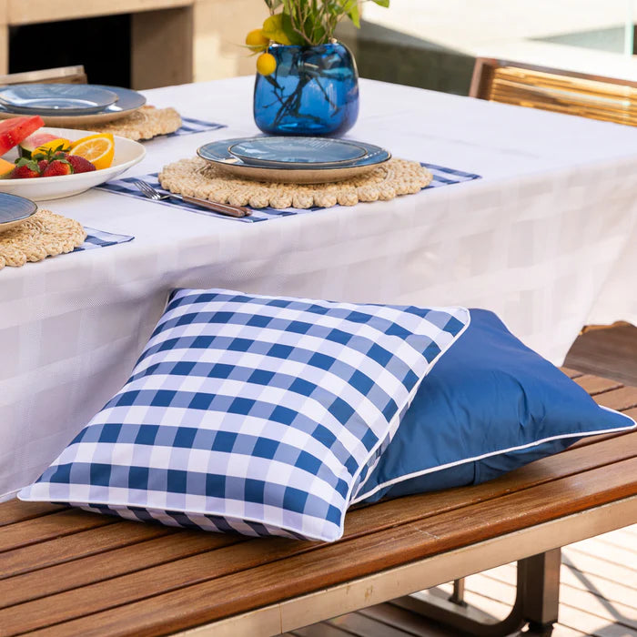 Blue and white checkered pillow on a wooden bench with a table and outdoor setting in the background.
