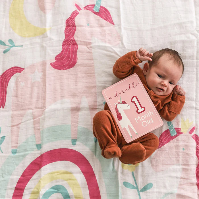 Baby in brown outfit holding a '1 Month Old' card on a colorful unicorn blanket